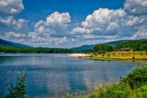 A serene lake surrounded by green hills and fluffy clouds, with a small beach and a house in the distance.