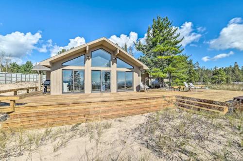A modern beach house with large windows, surrounded by trees and sandy terrain under a blue sky with clouds.