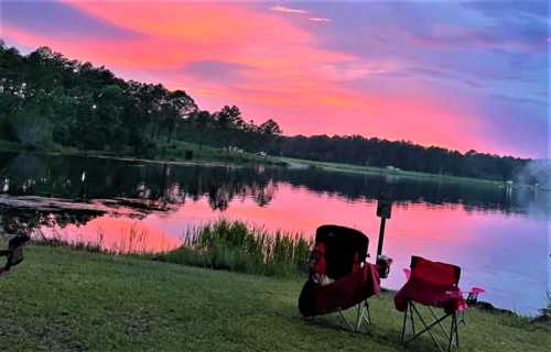 Two empty chairs by a calm lake at sunset, with vibrant pink and purple skies reflecting on the water.