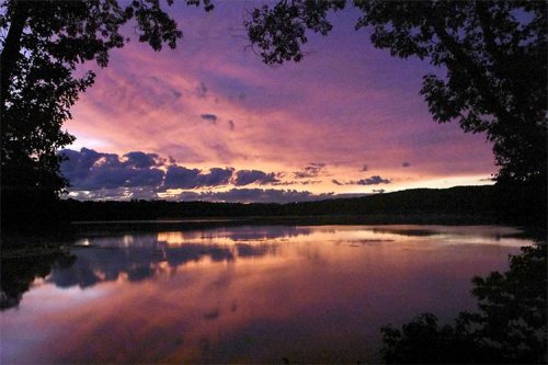 A serene lake at sunset, reflecting vibrant purple and orange hues in the sky, framed by trees on either side.