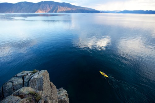 A lone kayaker paddles on calm waters, surrounded by mountains and a serene sky reflecting on the surface.