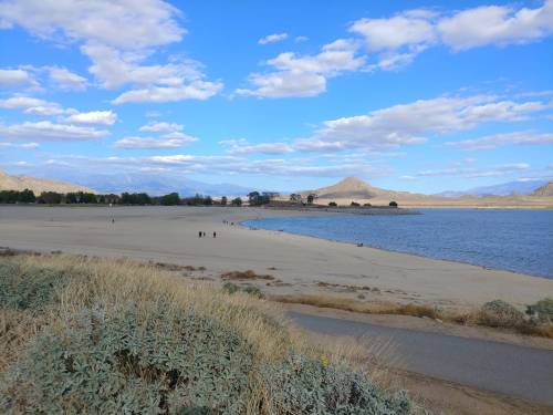 A scenic view of a sandy beach by a lake, with mountains in the background and a blue sky dotted with clouds.