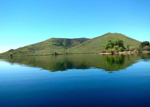 Serene landscape featuring green hills reflected in calm water under a clear blue sky.