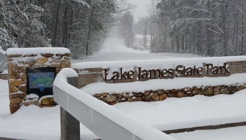 Snow-covered entrance sign for Lake James State Park, with a snowy road and trees in the background.