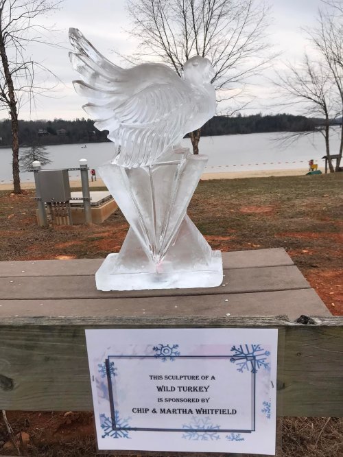 An ice sculpture of a wild turkey on a wooden stand by a lake, with a sign noting sponsorship by Chip & Martha Whitfield.