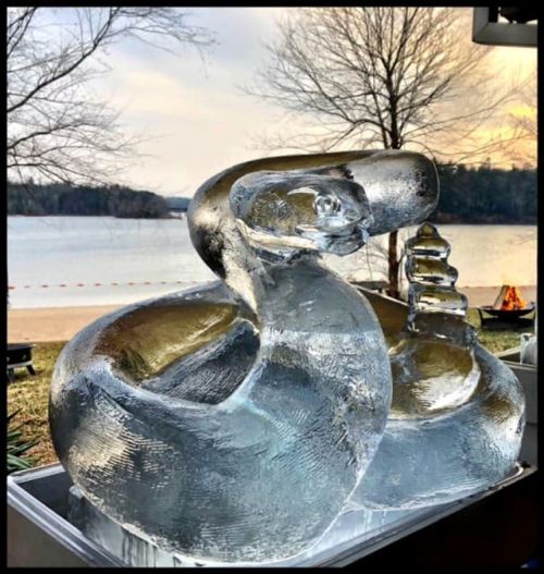 Two intricately carved ice sculptures of a swan and a pagoda, set against a serene lakeside backdrop.