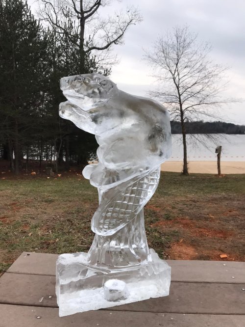 A large ice sculpture of a bear standing on its hind legs, set against a backdrop of trees and a lake.