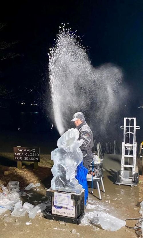 A man carves an ice sculpture, water spraying in the air, with a sign nearby stating "Swimming area closed for season."