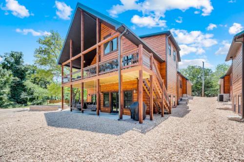 A modern wooden cabin with a balcony, surrounded by gravel and trees under a bright blue sky.