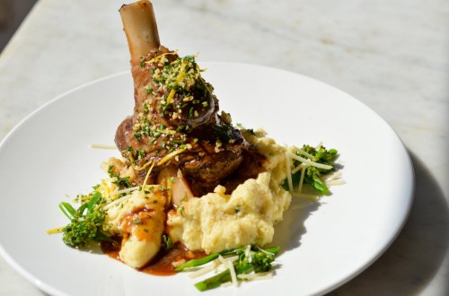 A plated dish featuring a braised lamb shank on a bed of creamy mashed potatoes, garnished with herbs and sauce.