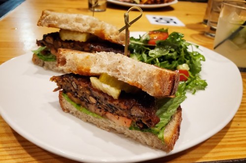 A close-up of a sandwich with layers of greens, pickles, and a crispy patty, served with a side salad on a white plate.
