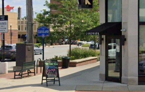 Street view featuring a sign for Cleveland, a restaurant sign, and a bench near a tree-lined sidewalk.