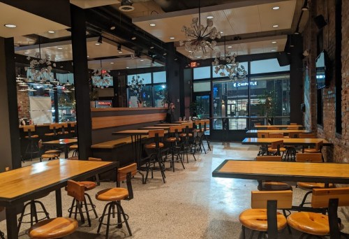 Interior of a modern restaurant with wooden tables, stools, and large windows, creating a spacious atmosphere.