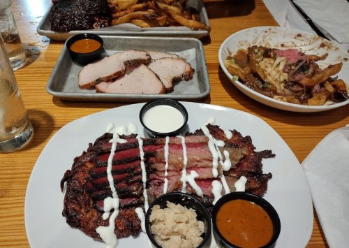 A plate of sliced barbecue meat with sauces, served alongside fries and a dish of loaded nachos.
