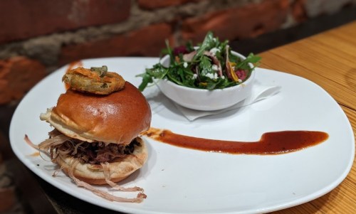 A pulled pork sandwich on a plate with a side salad and a drizzle of sauce.