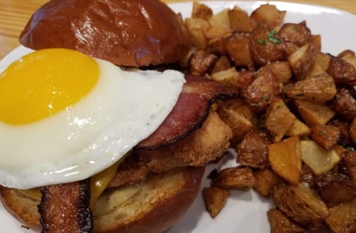 A sandwich with a fried egg, bacon, and chicken on a bun, served with crispy potato cubes on the side.
