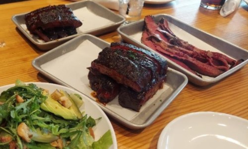 Three trays of barbecue meat, including brisket and ribs, alongside a fresh salad on a wooden table.