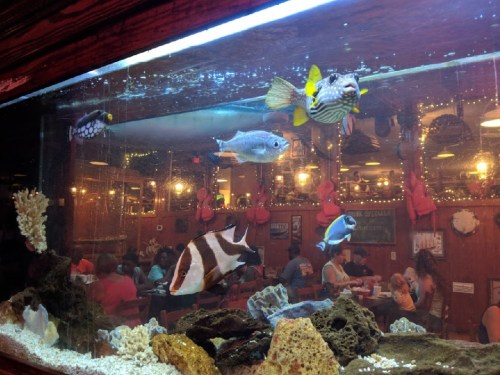 A vibrant aquarium with various fish swimming, set against a restaurant backdrop filled with diners.