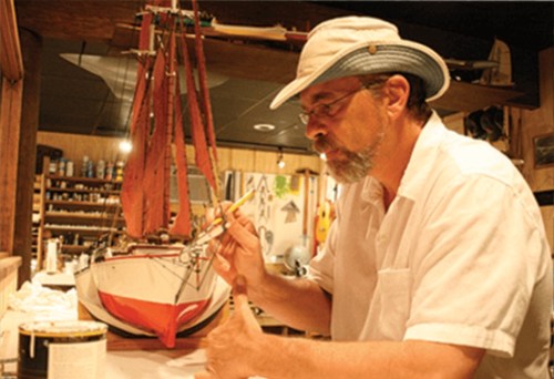 A man in a hat paints a model sailboat in a workshop filled with tools and materials.