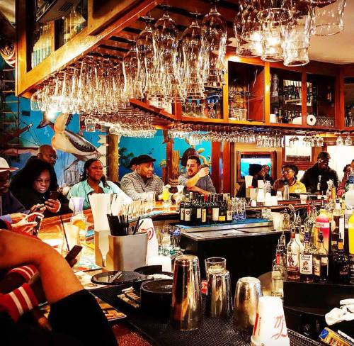 A lively bar scene with patrons chatting, drinks on the counter, and glasses hanging overhead. Colorful decor in the background.