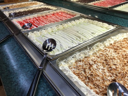 A variety of colorful toppings in trays, including shredded coconut, chocolate, and fruit, on a buffet table.