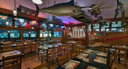 A rustic restaurant interior with wooden tables, chairs, and fishing-themed decor, including a large marlin hanging from the ceiling.