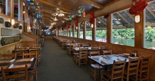 Interior of a rustic restaurant with wooden tables, chairs, and large windows overlooking a river.