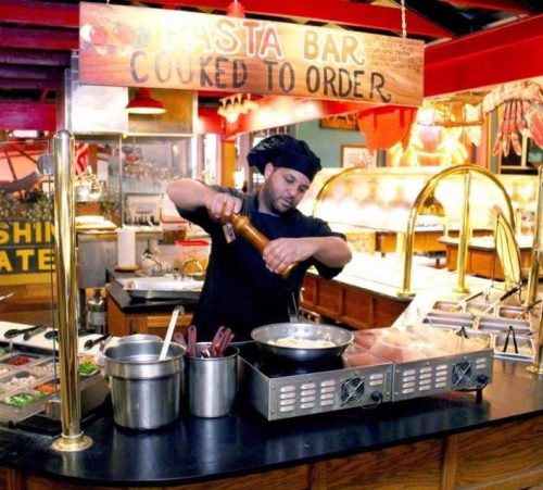 A chef prepares pasta at a bar, seasoning a dish with a pepper grinder in a vibrant restaurant setting.