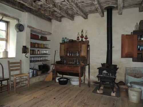 A rustic kitchen with wooden beams, a stove, shelves of jars, and a table, creating a cozy, vintage atmosphere.