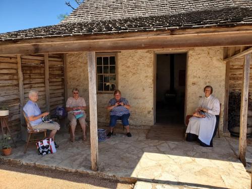 Four women sit outside a rustic building, engaged in knitting, with a stone wall and wooden structure in the background.