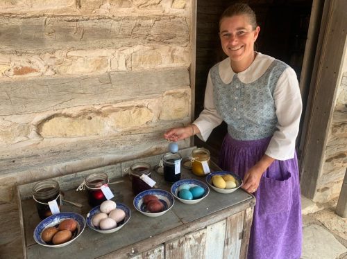 A woman in traditional dress stands by a table displaying dyed eggs in bowls and jars, smiling at the camera.