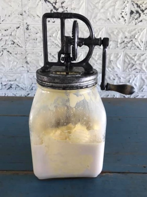 A vintage glass jar with a metal churn lid, containing a creamy mixture, set on a blue wooden table.