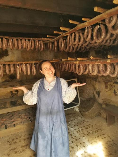 A woman in a blue apron smiles in a rustic room filled with hanging sausages.