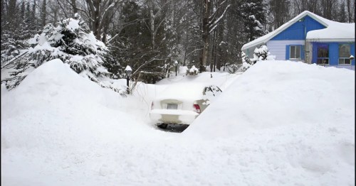 A car is partially buried in deep snow, surrounded by snowdrifts near a blue house in a winter landscape.