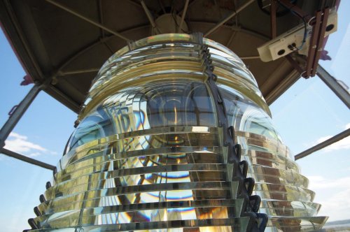 Close-up of a lighthouse lens, showcasing its intricate glass prisms and reflections under a clear blue sky.