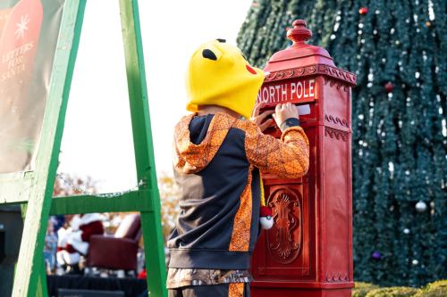 A child in a Pikachu hat drops a letter into a red North Pole mailbox near a decorated Christmas tree.
