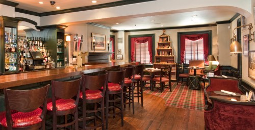 Cozy restaurant interior featuring a wooden bar, red upholstered chairs, and a bookshelf against a dark-paneled wall.
