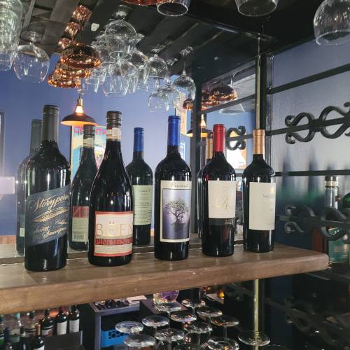 A wooden shelf displaying various wine bottles with hanging glasses in the background.