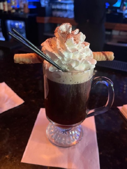 A glass of coffee topped with whipped cream and two rolled wafers, served on a bar counter.