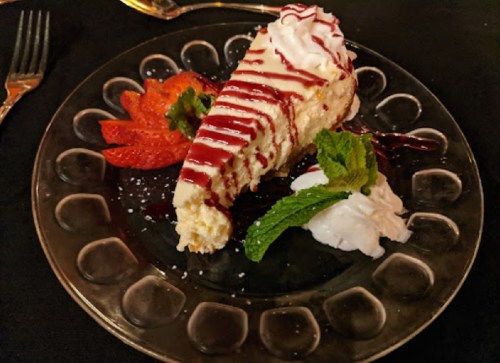 A slice of cheesecake drizzled with sauce, garnished with strawberries, mint, and whipped cream on a decorative plate.