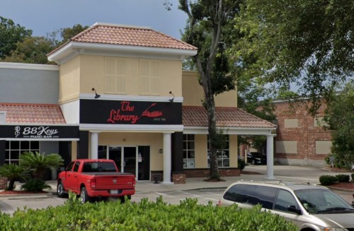A building with a sign reading "The Library," featuring a red truck parked in front and greenery in the foreground.