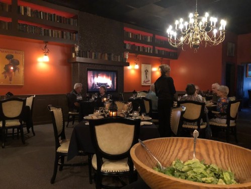 Cozy restaurant interior with a fireplace, diners at tables, and a server near a large salad bowl in the foreground.