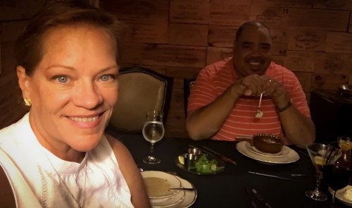 A woman smiles for a selfie with a man at a dining table, enjoying a meal in a cozy setting.