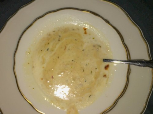 A bowl of creamy soup with a spoon, featuring a light color and herbs, served in an elegant dish.