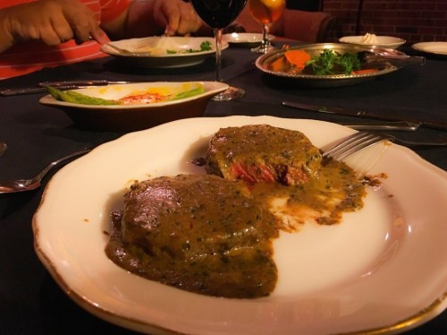 A plate with two pieces of meat covered in sauce, surrounded by a dark table setting and other dishes in the background.