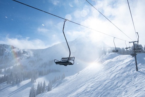 A ski lift chair hangs over a snowy mountain landscape under a bright, sunny sky.