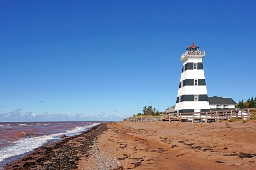 A black and white striped lighthouse stands by a sandy beach under a clear blue sky, with gentle waves lapping at the shore.