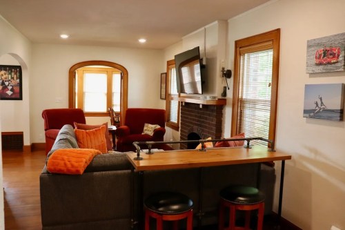 Cozy living room with red sofas, a wooden bar, and a TV above a fireplace, featuring large windows and warm lighting.