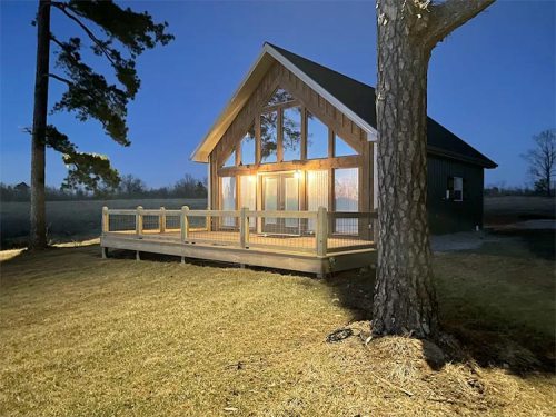 A cozy cabin with large windows and a deck, illuminated at dusk, surrounded by trees and open fields.