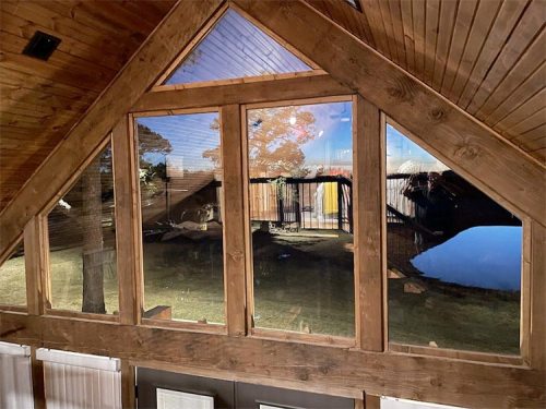 A wooden cabin interior with large windows reflecting a serene outdoor scene and a pond.